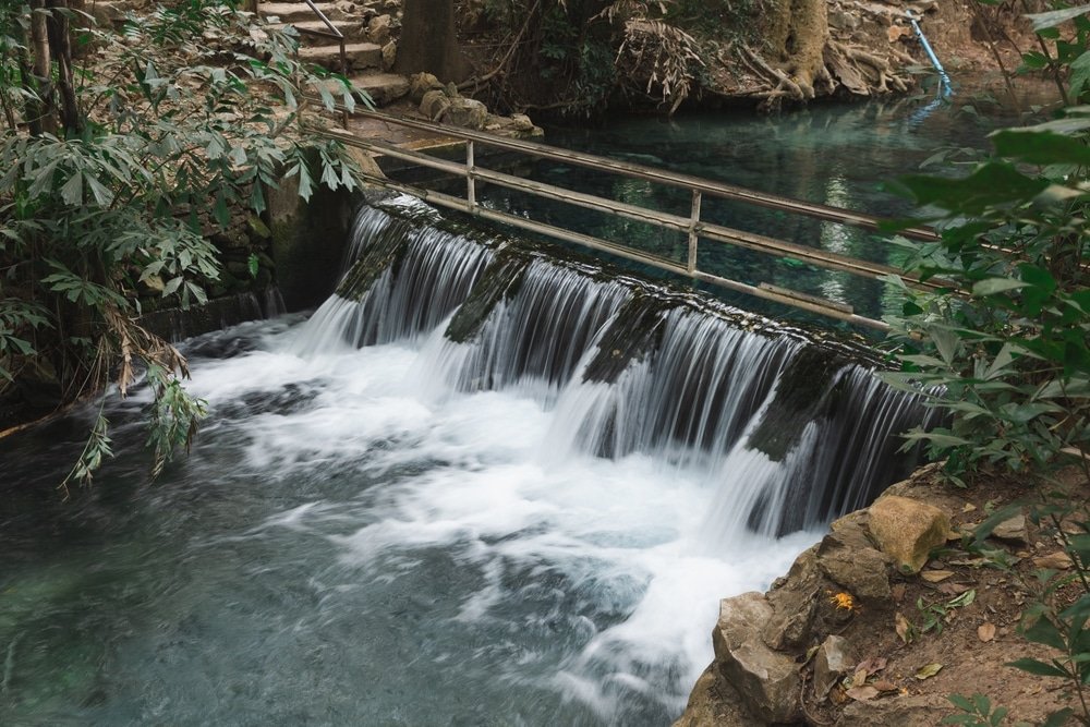 น้ำตกขนาดเล็กไหลผ่านสิ่งกีดขวางที่มนุษย์สร้างขึ้นพร้อมราวเหล็กในพื้นที่ป่า ล้อมรอบด้วยหินและพืชพรรณสีเขียว—จุดที่เหมาะสำหรับผู้ที่มองหาสถานที่ท่องเที่ยวเขาใหญ่ท่ามกลางความงดงามของธรรมชาติ.