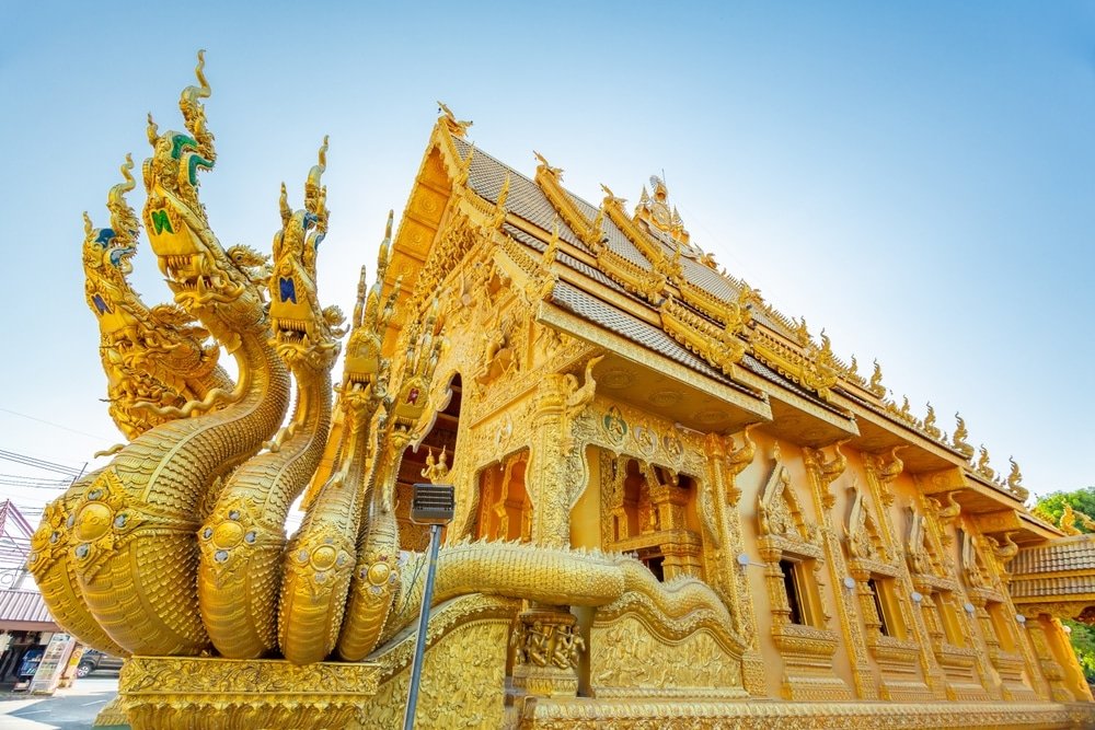 วัดพุทธทองคำที่มีลวดลายแกะสลักอันประณีตและรูปปั้นงูหลายหัวที่ทางเข้า ตั้งอยู่ท่ามกลางท้องฟ้าสีฟ้าใส—จุดที่ไม่ควรพลาดสำหรับผู้ที่มองหาสถานที่ท่องเที่ยวในน่าน.