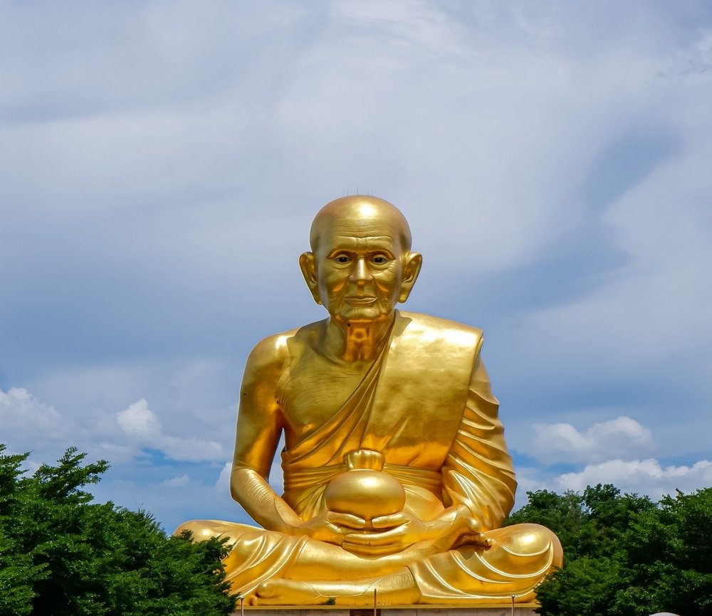 รูปปั้นพระพุทธรูปขนาดใหญ่สีทองของพระสงฆ์นั่งในจีวรแบบดั้งเดิม ตั้งอยู่ท่ามกลางต้นไม้และท้องฟ้าที่มีเมฆบางส่วน ทำให้เป็นสถานที่ท่องเที่ยวที่เงียบสงบในเขาใหญ่สำหรับผู้ที่มาเที่ยวเขาใหญ่.