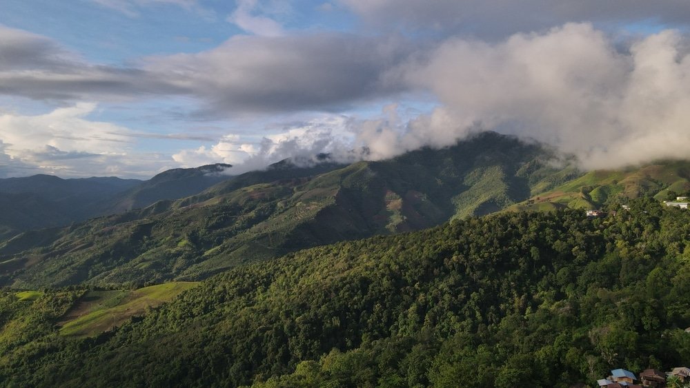 ภาพมุมสูงของภูเขาเขียวขจีปกคลุมด้วยป่าไม้ใน น่านที่เที่ยว มีเมฆกระจายและแสงแดดส่องผ่าน มองเห็นอาคารขนาดเล็กอยู่ไกลออกไป—เหมาะสำหรับผู้ที่ต้องการผจญภัยเที่ยวน่าน.