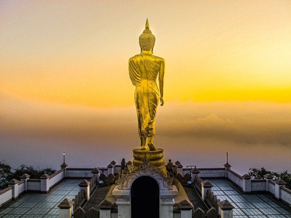 พระพุทธรูปทองคำขนาดใหญ่ตั้งอยู่บนแท่นสูง มองเห็นทิวทัศน์ที่ปกคลุมด้วยหมอกยามพระอาทิตย์ขึ้นหรือพระอาทิตย์ตกดิน โดยมีท้องฟ้าสีเหลืองส้มเป็นฉากหลัง—เป็นภาพอันเป็นเอกลักษณ์สำหรับผู้ที่มาเยือนหรือท่องเที่ยวในน่าน.