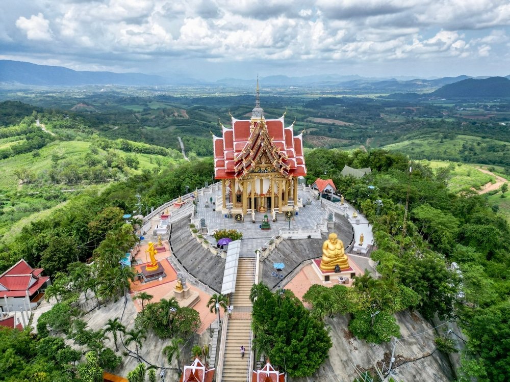 ภาพมุมสูงของวัดพุทธบนยอดเขาที่ล้อมรอบด้วยทิวทัศน์เขียวขจี พระพุทธรูปทองคำ และภูเขาไกล ๆ—สถานที่ท่องเที่ยวที่ไม่ควรพลาดในเขาใหญ่สำหรับผู้ที่มาเยือนภูมิภาคอันงดงามนี้.