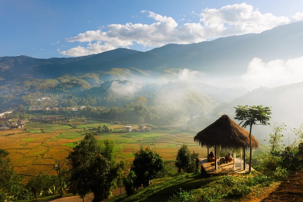 กระท่อมมุงจากตั้งอยู่เหนือทุ่งนาขั้นบันไดและเนินเขาที่ปกคลุมด้วยหมอก ภายใต้ท้องฟ้าสีครามที่มีเมฆลอยกระจายอยู่เป็นระยะ มีคนสองคนนั่งอยู่ภายในกระท่อม เพลิดเพลินกับทัศนียภาพอันงดงาม—เหมาะอย่างยิ่งสำหรับผู้ที่ต้องการท่องเที่ยวเมืองน่านและค้นพบสถานที่ท่องเที่ยวอันซ่อนเร้นในน่าน.