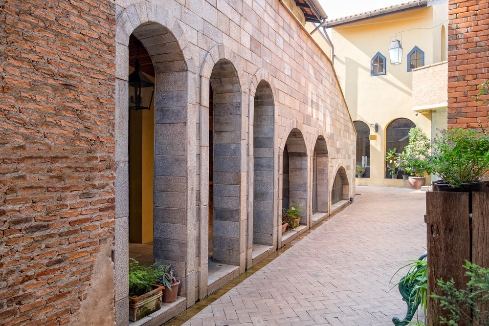 ตรอกแคบที่ปูด้วยอิฐขนาบข้างด้วยกำแพงอิฐและหินที่มีประตูโค้งและกระถางต้นไม้เรียงรายอยู่ตามด้านข้าง ชวนให้นึกถึง ที่เที่ยวเขาใหญ่ สำหรับผู้ที่มองหาสถานที่น่ารักๆ เพื่อสำรวจ.