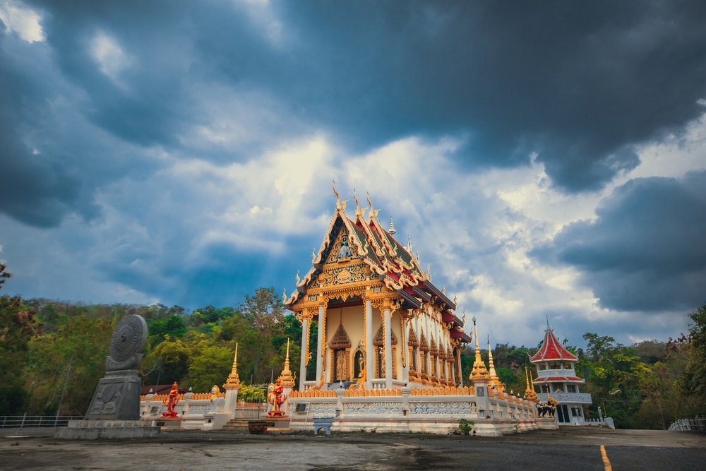 วัดไทยที่ประดับประดาอย่างวิจิตรพร้อมรายละเอียดหลังคาที่ซับซ้อนตั้งอยู่ใต้ท้องฟ้าที่มีเมฆครึ้มอย่างน่าทึ่ง ล้อมรอบด้วยพืชพรรณและรูปปั้น—สถานที่ท่องเที่ยวที่เงียบสงบในเขาใหญ่—ในขณะที่ศาลาเล็กๆ ตั้งอยู่อย่างเงียบสงบในฉากหลัง.