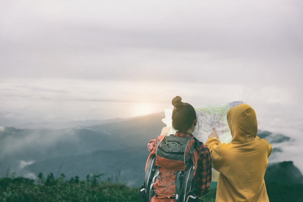 สองคนอยู่กลางแจ้งกำลังดูแผนที่ด้วยกัน โดยมีภูเขาและเมฆเป็นฉากหลัง—คนหนึ่งสะพายกระเป๋าเป้และสวมเสื้อเชิ้ตลายสก็อต อีกคนสวมเสื้อฮู้ดสีเหลือง—กำลังวางแผนการผจญภัยครั้งต่อไปที่ น่าน.