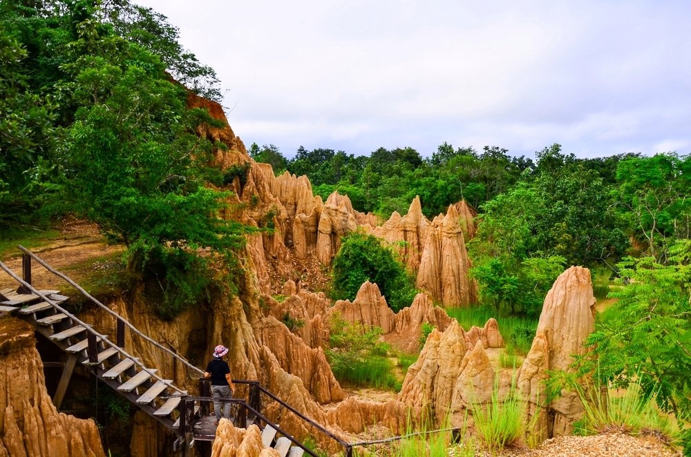 บุคคลหนึ่งยืนอยู่บนสะพานไม้ที่ทอดยาวออกไปเหนือการก่อตัวของทรายที่คมและถูกกัดเซาะ ล้อมรอบด้วยต้นไม้สีเขียวภายใต้ท้องฟ้าที่มีเมฆครึ้ม—หนึ่งในสถานที่ท่องเที่ยวที่น่าทึ่งของน่านสำหรับผู้รักธรรมชาติ.