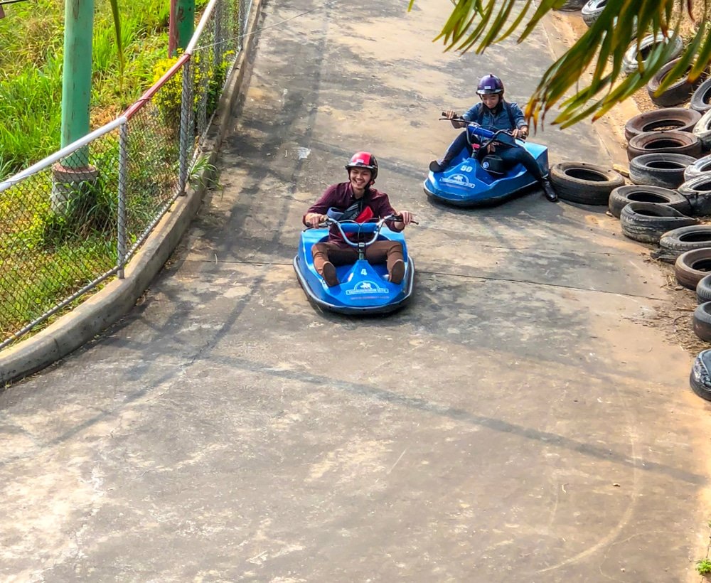 สองคนสวมหมวกกันน็อคขับรถโกคาร์ทสีน้ำเงินรอบสนามกลางแจ้งที่ปูพื้นด้วยคอนกรีต ล้อมรอบด้วยรั้วและยางรถยนต์ที่ซ้อนกันไว้ เหมาะสำหรับผู้ที่มองหาสถานที่ท่องเที่ยวเขาใหญ่หรือสถานที่ท่องเที่ยวที่น่าตื่นเต้นในเขาใหญ่.