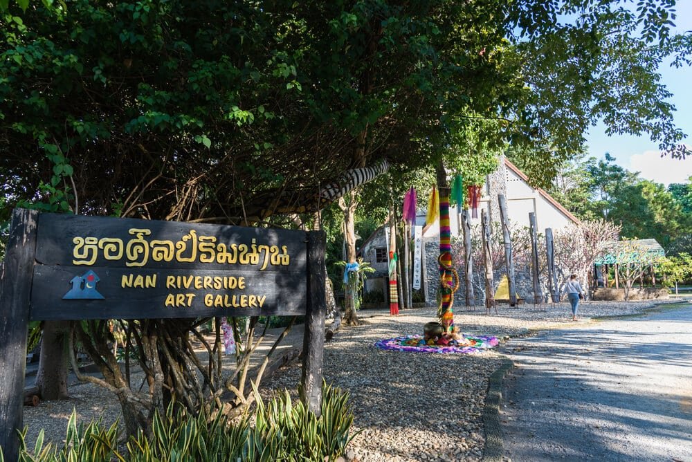 บริเวณทางเข้าของ Nan Riverside Art Gallery ซึ่งเป็นสถานที่ท่องเที่ยวอันดับต้น ๆ ในเมืองน่าน มีป้ายไม้, เสาหลากสี, ต้นไม้ และอาคารในพื้นหลังในวันที่แดดออก.
