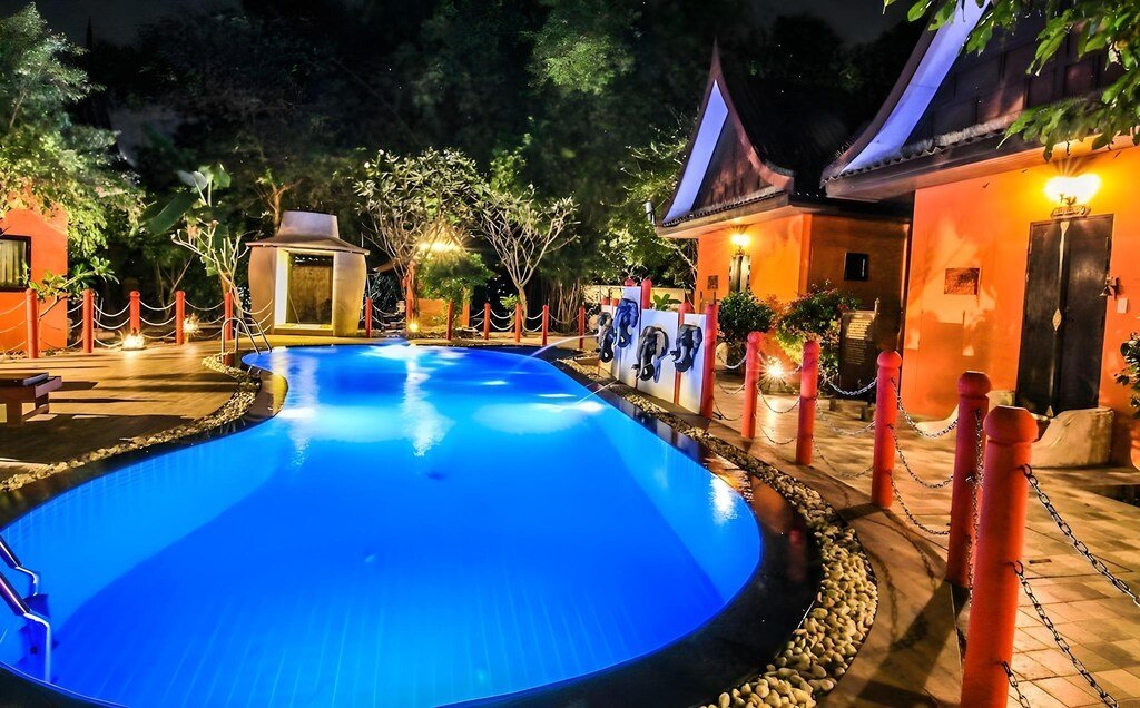 สระว่ายน้ำรูปไตพร้อมไฟสีน้ำเงินในยามค่ำคืน ล้อมรอบด้วยต้นไม้ เสาแดงพร้อมเชือก และอาคารหลังคาแหลม—เหมาะสำหรับค่ำคืนที่ผ่อนคลายที่ โรงแรมอยุธยา หรือที่พักริมน้ำอยุธยาสำหรับการพักผ่อนครั้งต่อไปของคุณ.