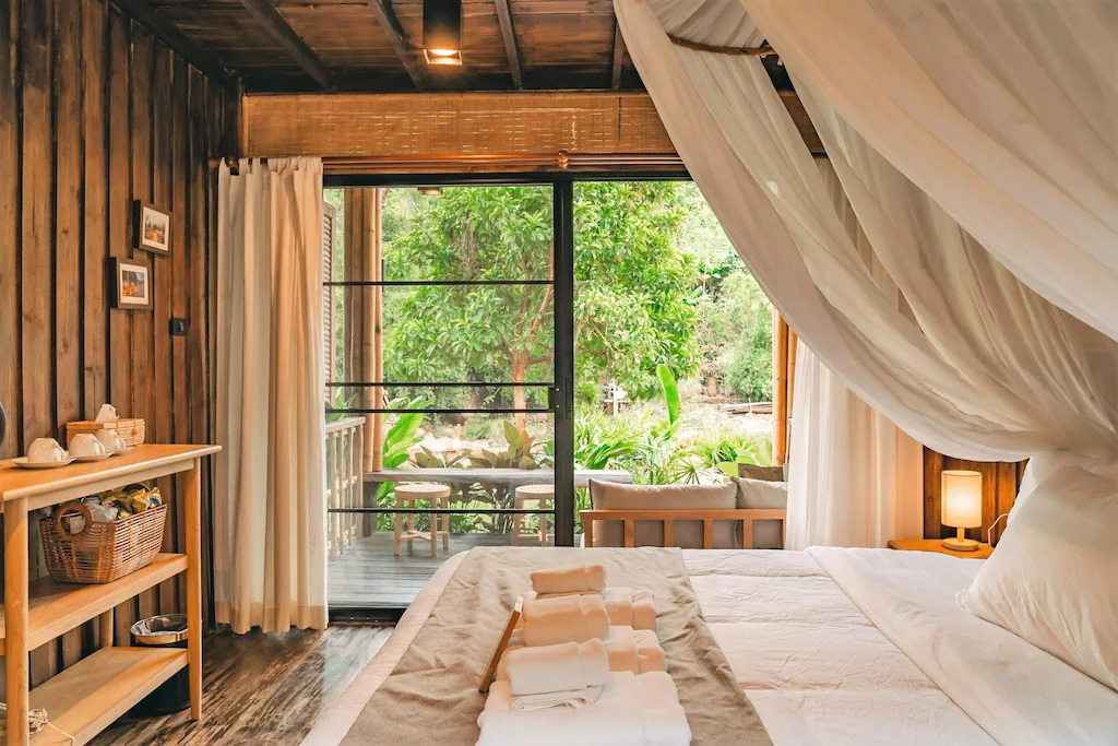 ห้องนอนไม้ที่อบอุ่นใน ที่พักน่านในเมือง พร้อมเตียงผ้าใบ, ผ้าเช็ดตัวบนผ้าห่ม, ชั้นวางเล็ก ๆ พร้อมชุดน้ำชาและตะกร้า, และหน้าต่างบานใหญ่ที่มองเห็นทิวทัศน์สีเขียวชอุ่ม.