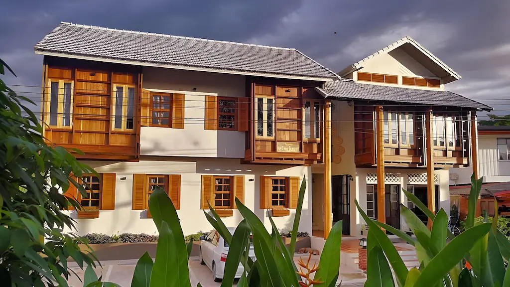 บ้านสองชั้นที่มีผนังสีขาวและกรอบหน้าต่างไม้ พร้อมที่จอดรถในร่มและพืชพรรณเขียวชอุ่มใต้ท้องฟ้าที่มีเมฆครึ้ม—เหมาะอย่างยิ่งสำหรับ ที่พักน่านในเมือง สำหรับผู้ที่ต้องการความสะดวกสบายท่ามกลางธรรมชาติ.