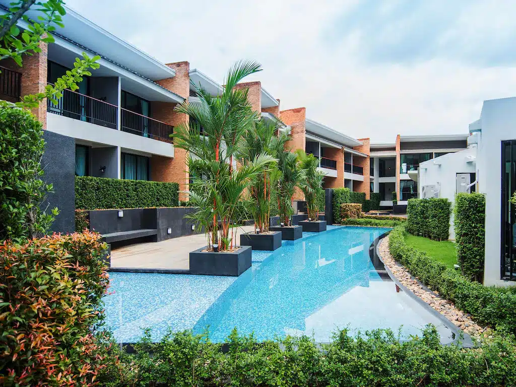 คอนโดมิเนียมสมัยใหม่พร้อมระเบียงที่มองเห็นสระว่ายน้ำกลางแจ้งยาว รายล้อมไปด้วยต้นไม้เขียวขจีและต้นปาล์มใต้ท้องฟ้าที่มีเมฆครึ้ม—เหมาะสำหรับการผ่อนคลายอย่างสมบูรณ์แบบหลังจากท่องเที่ยวสถานที่ท่องเที่ยวในอยุธยา.
