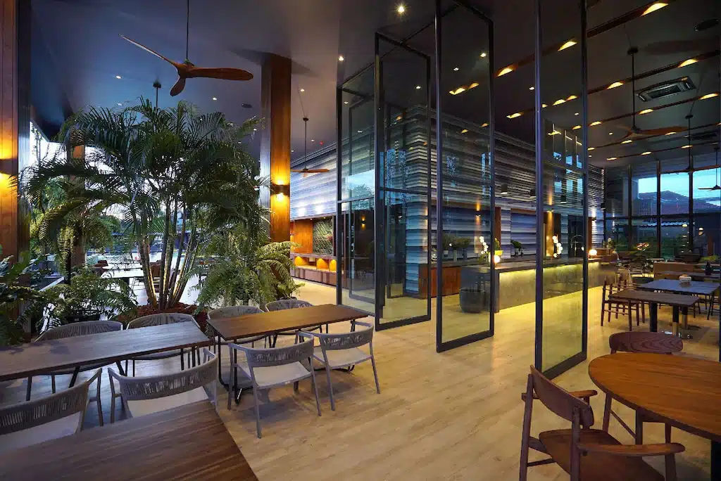 ร้านอาหารภายในอาคารสมัยใหม่ที่โรงแรมหัวหิน พร้อมโต๊ะและเก้าอี้ไม้, ผนังกั้นกระจกขนาดใหญ่, ต้นไม้ในร่มที่เขียวชอุ่ม, และแสงสว่างอบอุ่น; บริเวณสระว่ายน้ำกลางแจ้งของที่พักติดทะเลหัวหินที่สามารถมองเห็นได้ผ่านหน้าต่าง.