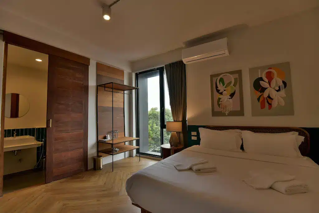 ห้องพักโรงแรมอยุธยาสมัยใหม่พร้อมเตียงคู่ ผ้าเช็ดตัวสองผืนพับไว้ ศิลปะบนผนังเหนือเตียง หน้าต่างบานใหญ่ ชั้นวางของแบบเปิดโล่ง และห้องน้ำในตัวพร้อมประตูบานเลื่อนไม้.