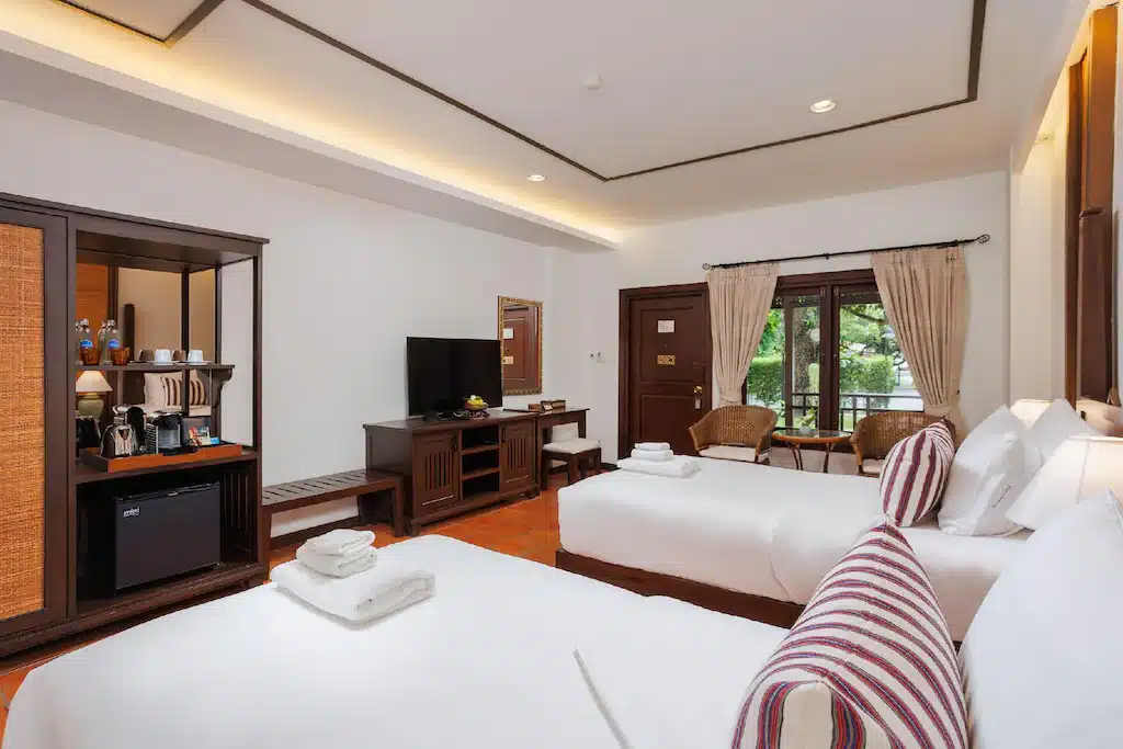ห้องพักโรงแรมพร้อมเตียงสองเตียง โทรทัศน์บนตู้ไม้ มินิบาร์พร้อมอุปกรณ์ชงกาแฟ เก้าอี้สองตัวริมหน้าต่าง และแสงไฟอบอุ่น—เหมาะสำหรับการพักผ่อนหลังจากสำรวจสถานที่ท่องเที่ยวเชียงใหม่.