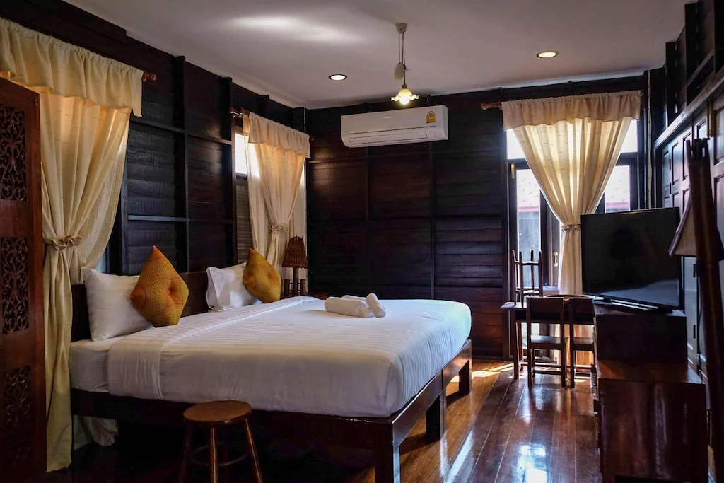 ห้องพักโรงแรมที่ โรงแรมอยุธยา พร้อมผนังไม้สีเข้ม เตียงใหญ่พร้อมผ้าปูที่นอนสีขาว หมอนประดับสีทอง โทรทัศน์ เฟอร์นิเจอร์ไม้ และหน้าต่างพร้อมผ้าม่านสีครีมที่เปิดให้แสงแดดส่องเข้ามา.