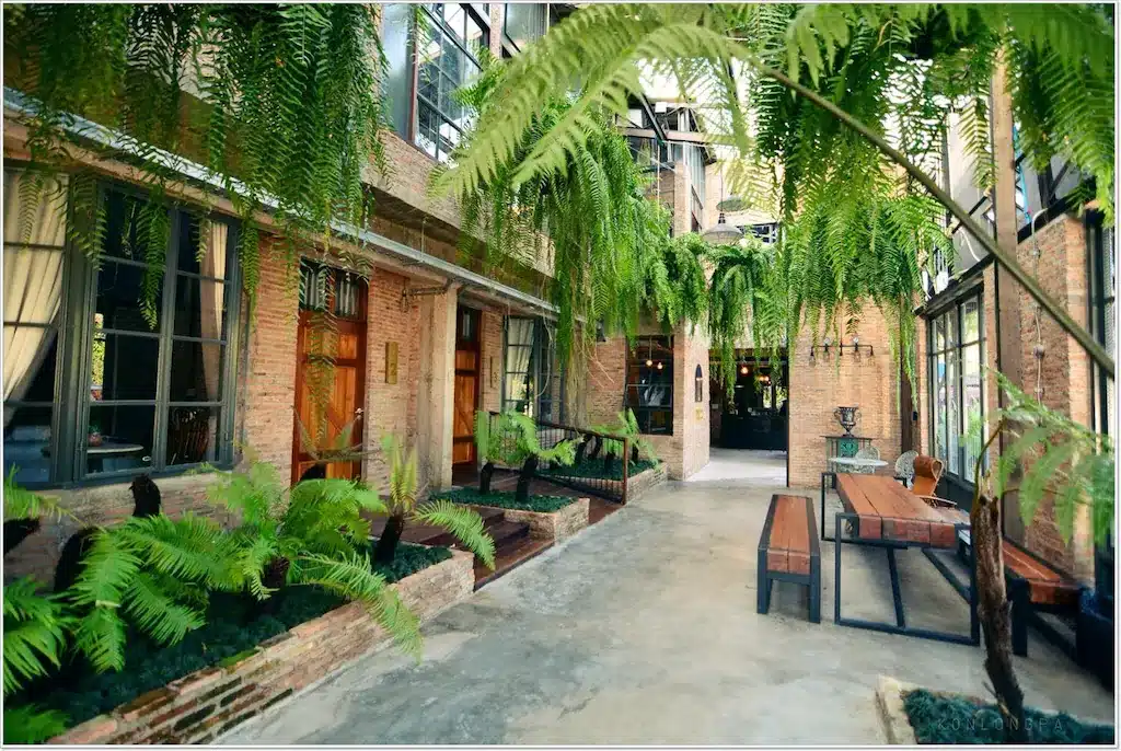 ลานกลางแจ้งในร่มกว้างขวางพร้อมผนังอิฐ หน้าต่างบานใหญ่ เฟิร์นแขวน ประตูไม้ ม้านั่ง และโต๊ะไม้ยาวบนพื้นคอนกรีต—เหมาะสำหรับการพักผ่อนที่ ที่พักน่านในเมือง.