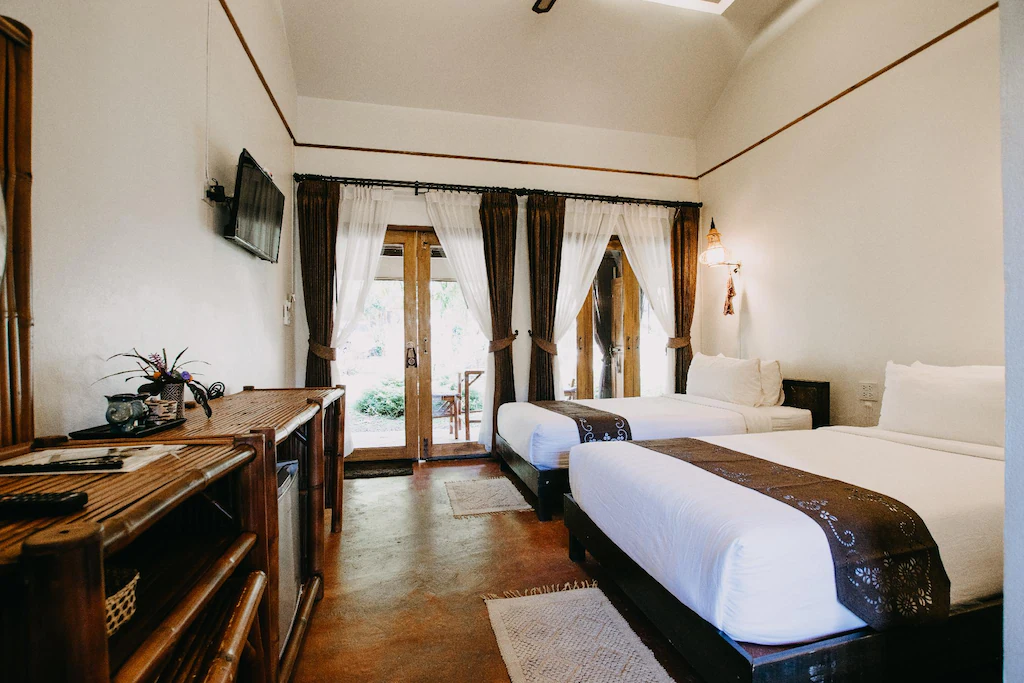 ห้องพักโรงแรมพร้อมเตียงคู่สองเตียง ผ้าปูที่นอนสีขาว โทรทัศน์ติดผนัง เฟอร์นิเจอร์หวาย และประตูกระจกที่เปิดออกสู่ลานระเบียงพร้อมพื้นที่สีเขียวภายนอก—เหมาะสำหรับการเข้าพักของคุณที่ ที่พักน่าน.