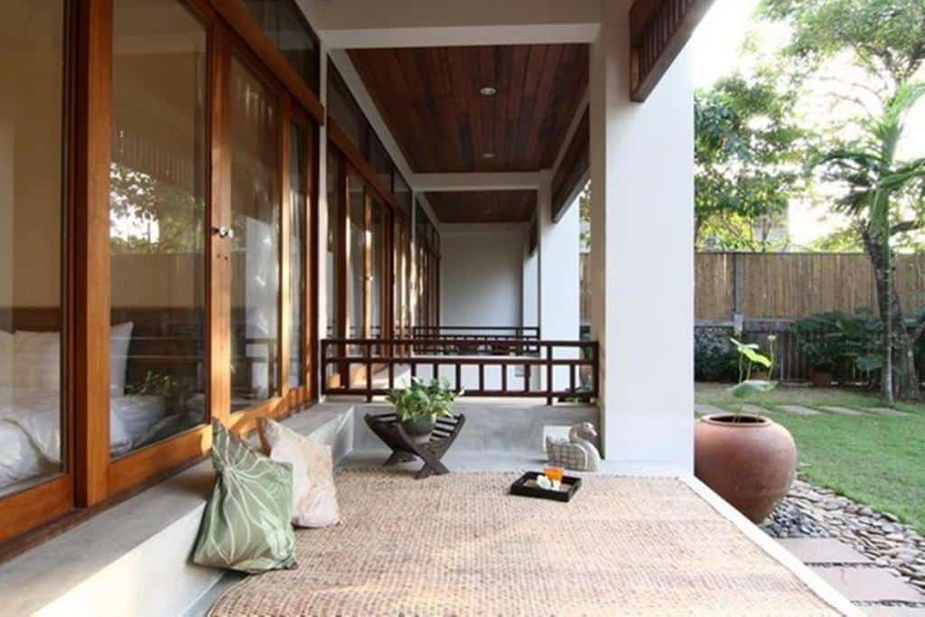 ระเบียงที่มีหลังคาคลุม พร้อมเสื่อสาน หมอนอิง โต๊ะเล็ก และต้นไม้ประดับอยู่ข้างสนามหญ้าและกระถางดินเผาขนาดใหญ่ถัดจากประตูกระจกบานเลื่อน—เหมาะอย่างยิ่งสำหรับการพักผ่อนหลังจากเที่ยวชมสถานที่ท่องเที่ยวในอยุธยา.