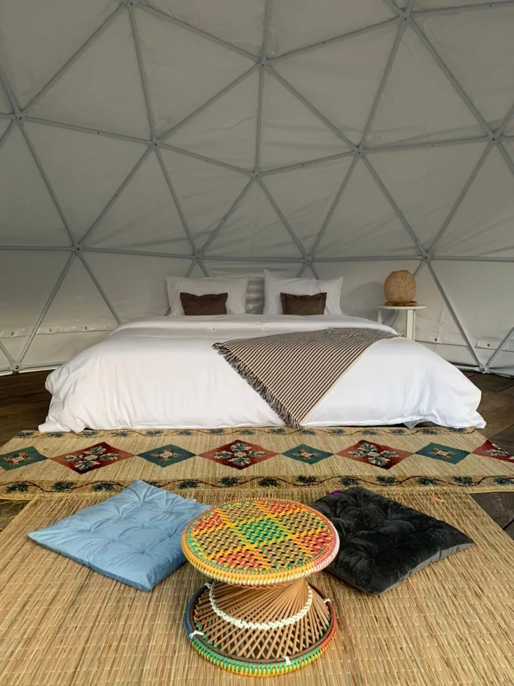 ห้องทรงโดมที่มีเตียงขนาดใหญ่ ผ้าห่มลายทาง พรมลาย และเบาะรองนั่งสองใบบนพื้น มอบที่พักแสนสบายในแบบ ที่พักปัว พร้อมโต๊ะกลมสานขนาดเล็กวางอยู่หน้าเตียง.