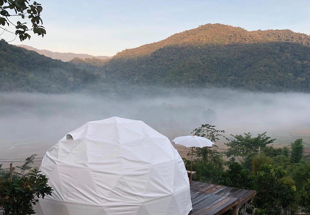 โดมจีโอเดสิกสีขาวตั้งอยู่บนดาดฟ้าไม้ มองเห็นวิวเนินเขาที่ปกคลุมด้วยหมอกและพืชพรรณเขียวขจีภายใต้ท้องฟ้าแจ่มใส เหมาะสำหรับผู้ที่มองหาที่พักบ่อเกลือหรือที่พักน่านที่ไม่เหมือนใครท่ามกลางธรรมชาติ.