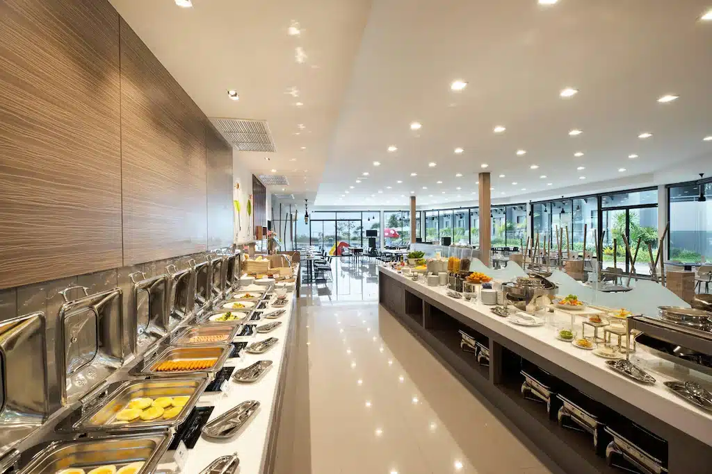 ร้านอาหารบุฟเฟ่ต์สไตล์ที่มีเคาน์เตอร์ยาวโชว์อาหารร้อนและเย็นหลากหลายชนิด ตกแต่งสไตล์โมเดิร์น พร้อมหน้าต่างขนาดใหญ่ที่เปิดให้แสงธรรมชาติส่องผ่าน—เหมาะสำหรับแขกที่พักอยู่ที่ ที่พักหัวหิน หรือกำลังค้นหา ที่พักหัวหินราคาถูก ในบริเวณใกล้เคียง.