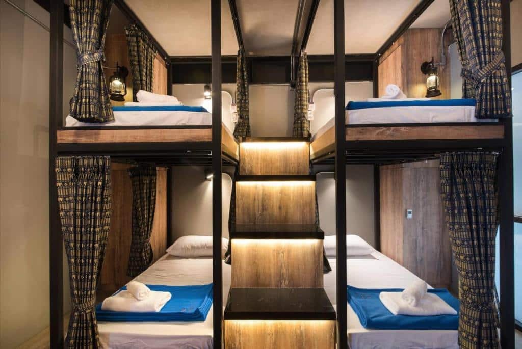 ห้องพักโฮสเทลสมัยใหม่พร้อมเตียงสองชั้นสี่เตียง แต่ละเตียงมีผ้าปูที่นอนสีน้ำเงินและผ้าเช็ดตัวม้วน ตกแต่งด้วยไม้ ม่านบังตาเพื่อความเป็นส่วนตัว และบันไดที่มีแสงไฟ—เหมาะสำหรับนักเดินทางที่กำลังมองหา ที่พักอยุธยา ที่สะดวกสบายและมีสไตล์.