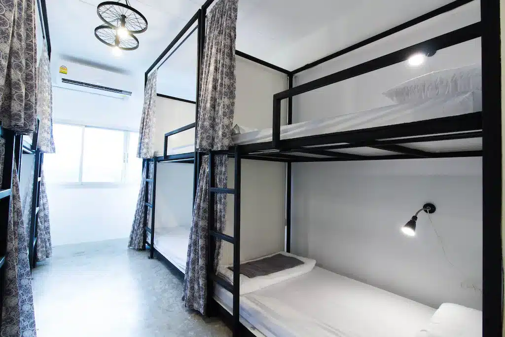 ห้องพักในโฮสเทลพร้อมเตียงสองชั้นสี่เตียง แต่ละเตียงมีม่านสำหรับความเป็นส่วนตัว เครื่องนอนสีขาว และไฟอ่านหนังสือส่วนตัว เหมาะสำหรับนักเดินทางที่ต้องการความสะดวกสบายในโรงแรมอยุธยา พร้อมหน้าต่างที่ให้แสงธรรมชาติ.