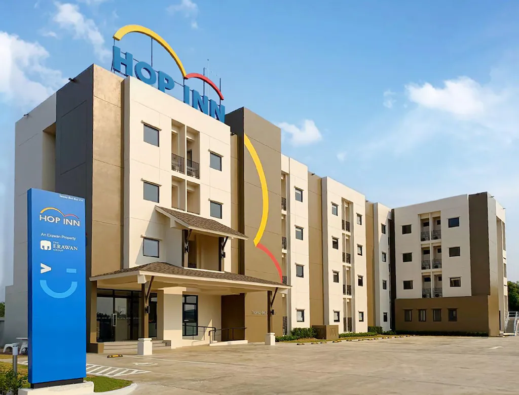 โรงแรมสมัยใหม่สี่ชั้นพร้อมป้าย "HOP INN" บนหลังคาและป้ายทางเข้าสีน้ำเงินด้านหน้า เหมาะสำหรับผู้ที่มองหาที่พักในปัวหรือที่พักในน่าน ท่ามกลางท้องฟ้าสีฟ้าใส.
