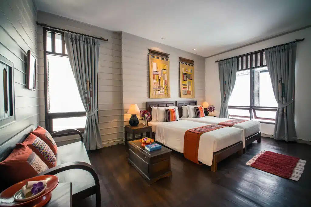 ห้องพักโรงแรมที่จัดเรียงอย่างเรียบร้อยที่ ที่พักบ่อเกลือ พร้อมเตียงคู่สองเตียง พื้นไม้ ม้านั่ง โต๊ะเป็นหีบไม้ โคมไฟ และหน้าต่างบานใหญ่พร้อมผ้าม่านสีเทาที่ปล่อยให้แสงธรรมชาติส่องเข้ามา.