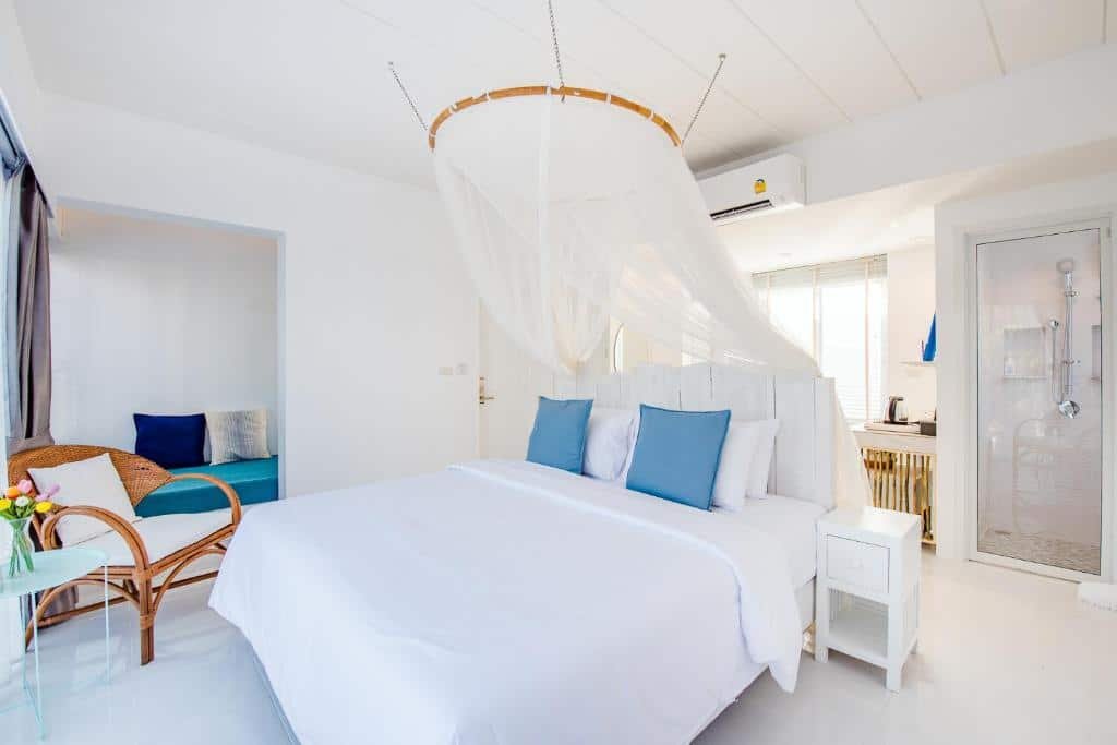 ห้องนอนสว่างพร้อมเตียงสีขาว หมอนอิงสีน้ำเงิน ผ้าม่านกันยุงแบบเต็นท์ เก้าอี้หวาย และพื้นที่นั่งเล่น เพลิดเพลินกับที่พักแสนสบายในหัวหินพร้อมเครื่องปรับอากาศและห้องครัวขนาดเล็ก—เหมาะสำหรับการพักผ่อนใกล้ทะเล.