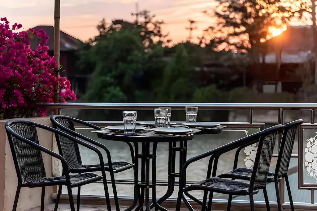 โต๊ะกลางแจ้งขนาดเล็กพร้อมเก้าอี้สีดำสี่ตัว แก้วน้ำ และจานวางอยู่บนระเบียงยามพระอาทิตย์ตกดิน สร้างบรรยากาศอันเงียบสงบของอยุธยาที่เที่ยว โดยมีดอกไม้และต้นไม้เป็นฉากหลัง.