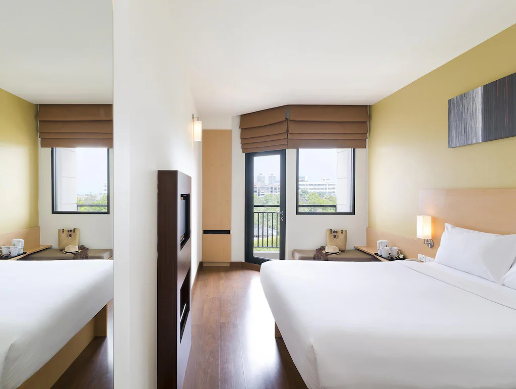 ห้องพักโรงแรมสมัยใหม่ใน โรงแรมหัวหิน พร้อมเตียงคู่ที่จัดไว้อย่างเรียบร้อย พื้นไม้สีอ่อน กระจกบานใหญ่ หน้าต่างสองบานพร้อมม่านสีน้ำตาล และทีวีติดผนัง—เหมาะสำหรับผู้ที่มองหา ที่พักหัวหินราคาถูก.