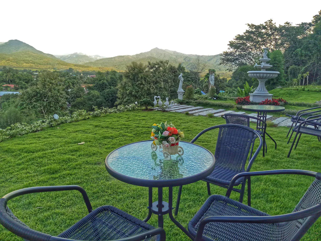 โต๊ะกระจกพร้อมการจัดดอกไม้และเก้าอี้ว่างบนสนามหญ้า มองเห็นสวนที่มีรูปปั้นและน้ำพุ ภูเขาเป็นฉากหลัง—เหมาะสำหรับการพักผ่อนที่ ที่พักบ่อเกลือ หรือ ที่พักปัว ที่คุณชื่นชอบ.