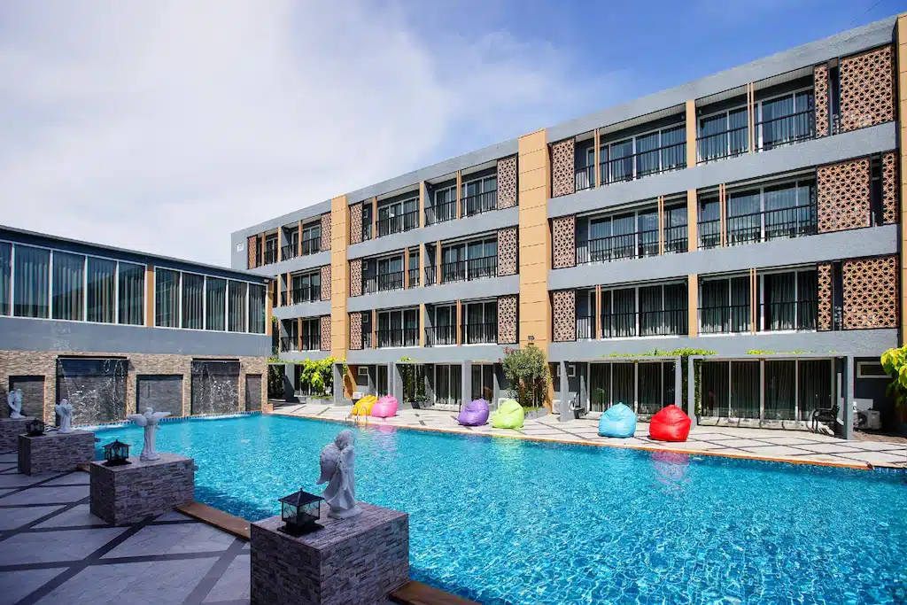 โรงแรมหัวหินสมัยใหม่ 3 ชั้น พร้อมสระว่ายน้ำกลางแจ้งขนาดใหญ่ เบาะนั่งรูปถั่วสีสันสดใสริมสระ รวมถึงรูปปั้นและน้ำตก—เหมาะสำหรับประสบการณ์ที่พักหัวหินราคาประหยัดที่ทั้งมีสไตล์และคุ้มค่า.