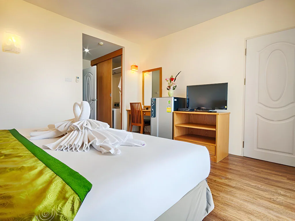 ห้องพักโรงแรมที่ โรงแรมอยุธยา พร้อมเตียงที่จัดไว้อย่างเรียบร้อย ผ้าเช็ดตัวพับเป็นรูปนกยูง โทรทัศน์บนขาตั้งไม้ ตู้เย็นขนาดเล็ก โต๊ะพร้อมเก้าอี้ และพื้นไม้—ความสะดวกสบายที่สมบูรณ์แบบสำหรับการเข้าพักของคุณในอยุธยา.