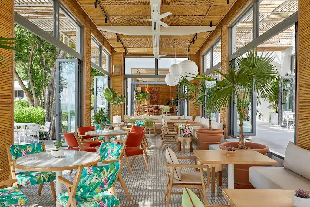 ร้านอาหารสมัยใหม่ที่มีแสงแดดส่องถึงพร้อมหน้าต่างบานใหญ่ ตกแต่งด้วยไม้ พืชเขตร้อน โต๊ะหินอ่อนและไม้ และเก้าอี้สีสันสดใส—เหมาะสำหรับแขกที่กำลังมองหาที่พักหัวหินราคาถูกหรือโรงแรมหัวหินใกล้เคียง.