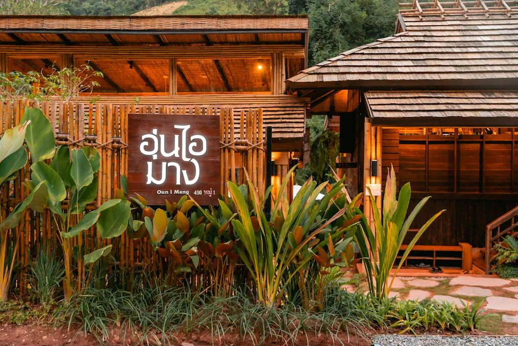อาคารไม้หลังคาไม้ระแนงพร้อมต้นไม้เขียวชอุ่มด้านหน้า ป้ายที่เขียนด้วยอักษรไทยใกล้ทางเข้าบ่งบอกถึงเสน่ห์ของที่พักแท้ในเมืองน่าน.