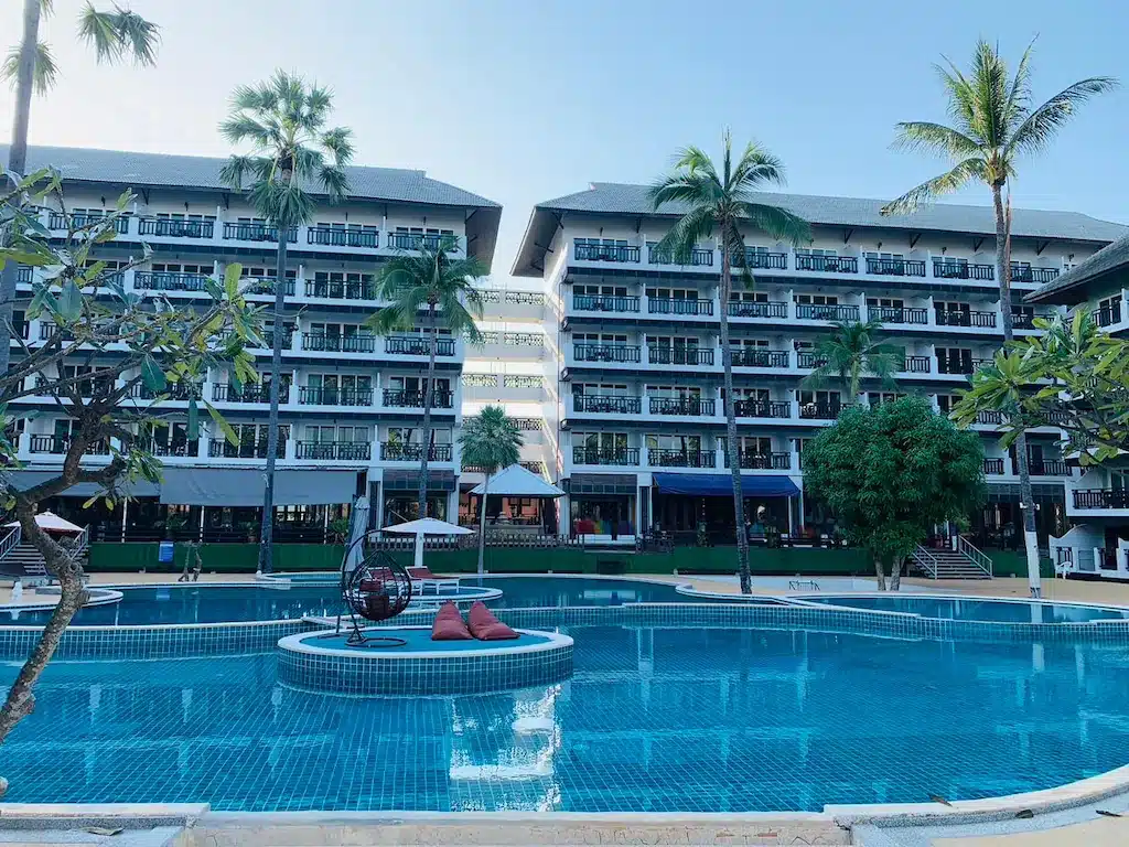 สระว่ายน้ำกลางแจ้งขนาดใหญ่พร้อมเกาะกลางรูปวงกลมและเก้าอี้อาบแดดตั้งอยู่ด้านหน้าโรงแรมหลายชั้นที่เรียงรายไปด้วยระเบียงและล้อมรอบด้วยต้นปาล์ม—เหมาะสำหรับผู้ที่มองหาที่พักหัวหินราคาถูกใกล้ทะเล.