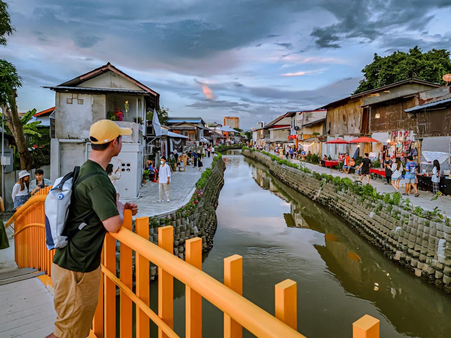 บุคคลหนึ่งสะพายกระเป๋าเป้ยืนอยู่บนสะพานสีเหลือง มองเห็นคลองที่เรียงรายไปด้วยบ้านเรือนและแผงขายของตลาดภายใต้ท้องฟ้าที่มีเมฆครึ้ม กำลังบันทึกเสน่ห์ของการเที่ยวเชียงใหม่และสถานที่ท่องเที่ยวเชียงใหม่ที่ไม่เหมือนใคร.