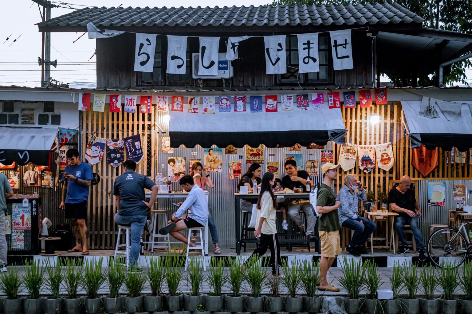 ผู้คนรับประทานอาหารและสังสรรค์กันนอกร้านอาหารสไตล์ญี่ปุ่นในเชียงใหม่ ซึ่งตกแต่งด้วยแบนเนอร์และโปสเตอร์สีสันสดใส—สถานที่ยอดนิยมสำหรับ เที่ยวเชียงใหม่ โดยมีต้นไม้และจักรยานอยู่ด้านหน้า.