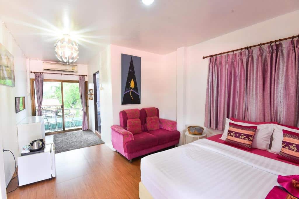 ห้องนอนสว่างไสวที่โรงแรมอยุธยา พร้อมเตียงคู่ ผ้าม่านสีชมพู โซฟาสีชมพู ภาพศิลปะติดผนัง ตู้เย็นขนาดเล็ก และระเบียงที่สามารถมองเห็นวิวสีเขียวชอุ่ม.