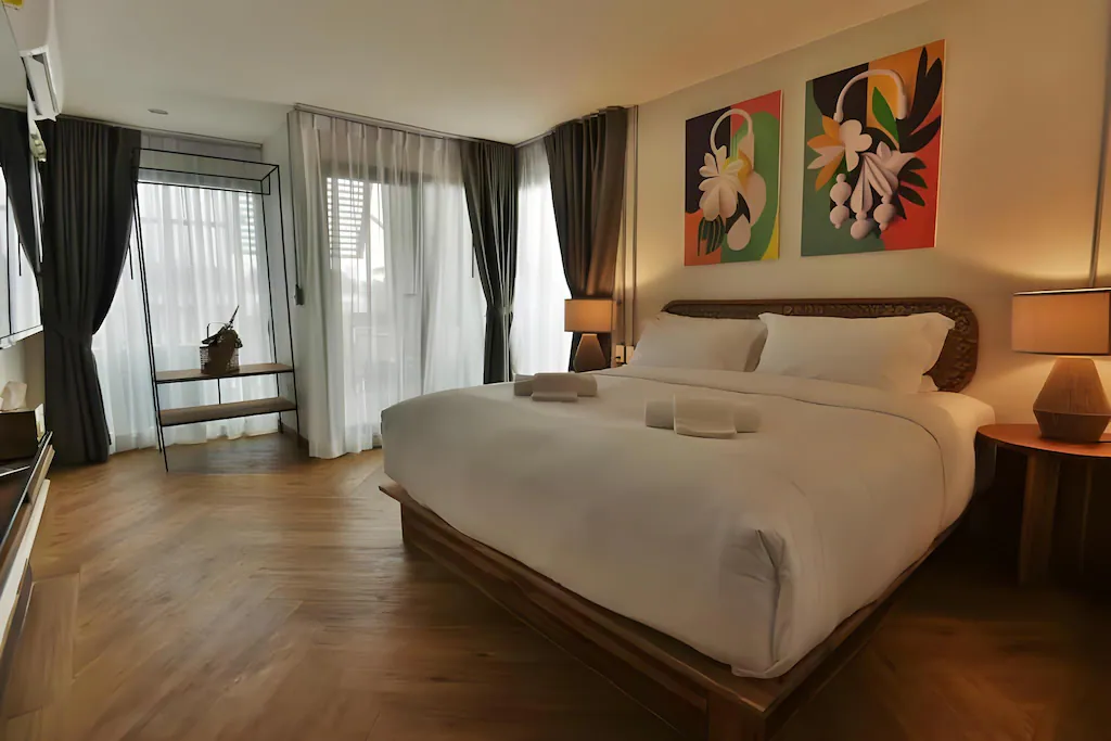 ห้องพักโรงแรมสมัยใหม่ที่ ที่พักอยุธยา พร้อมเตียงขนาดใหญ่ โคมไฟข้างเตียงสองดวง โต๊ะทำงานริมหน้าต่าง และภาพวาดดอกไม้สีสันสดใสสองภาพเหนือหัวเตียง เหมาะสำหรับการเข้าพักของคุณในอยุธยา.