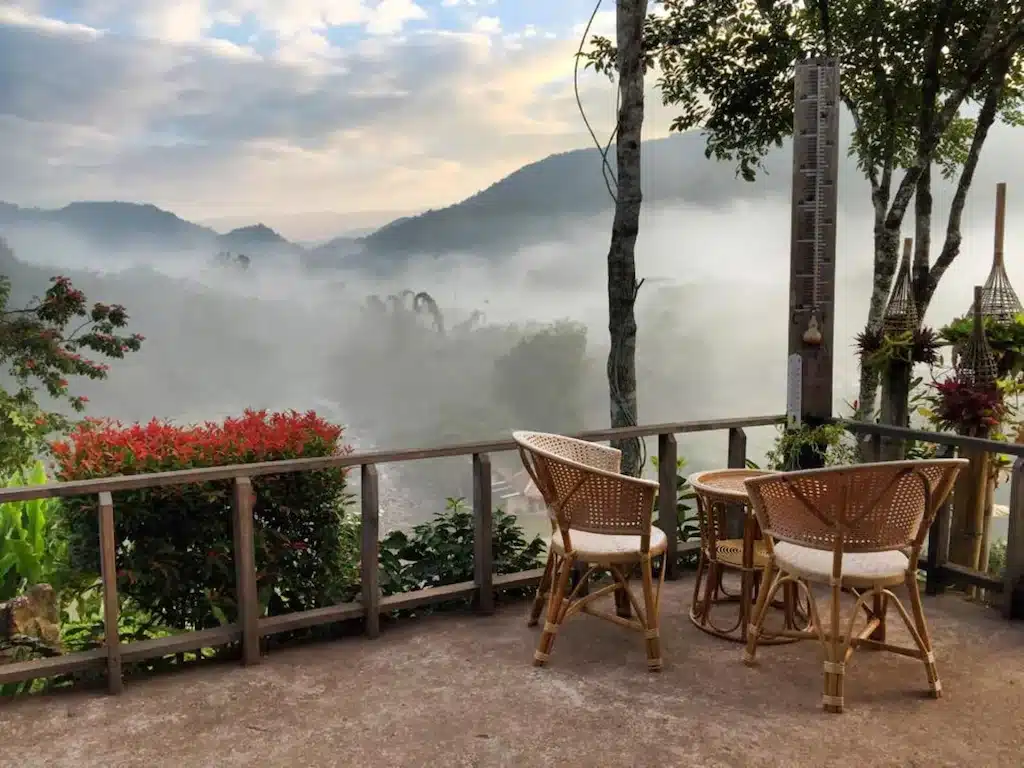 ลานระเบียงกลางแจ้งพร้อมโต๊ะกลมและเก้าอี้หวายสามตัว มองเห็นหุบเขาที่มีหมอกใน น่าน เหมาะสำหรับการพักผ่อนที่ ที่พักน่าน ท่ามกลางภูเขาและต้นไม้ภายใต้ท้องฟ้าที่มีเมฆปกคลุม.