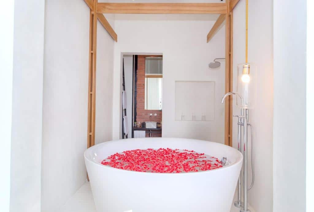 อ่างอาบน้ำสีขาวขนาดใหญ่ที่เต็มไปด้วยน้ำและกลีบกุหลาบตั้งอยู่ในห้องน้ำสไตล์มินิมอลที่มีผนังสีขาวและตกแต่งด้วยไม้ สร้างบรรยากาศผ่อนคลายที่ชวนให้นึกถึงการเข้าพักในบูติกโฮเทลใกล้แหล่งท่องเที่ยวอยุธยา.