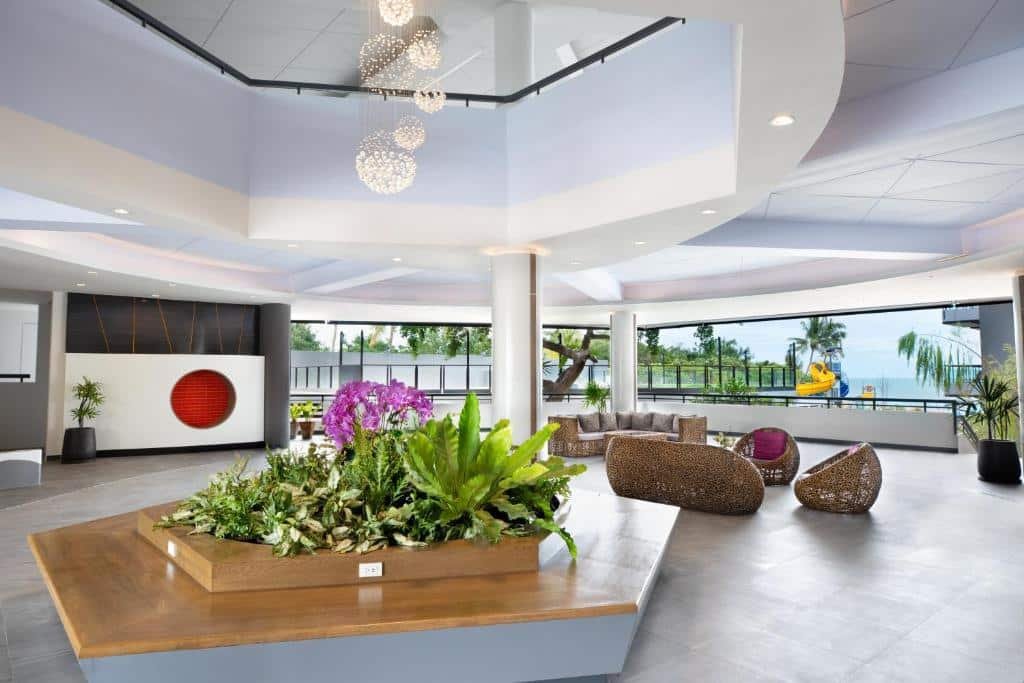 ล็อบบี้โรงแรมสมัยใหม่ที่ โรงแรมหัวหิน โดดเด่นด้วยกระถางต้นไม้ขนาดใหญ่ในร่ม, ที่นั่งหวาย, การจัดดอกกล้วยไม้, และหน้าต่างสูงจากพื้นจรดเพดานที่มองเห็นสระว่ายน้ำและต้นปาล์มด้านนอก—ประสบการณ์ที่พักติดทะเลหัวหินที่มีสไตล์.