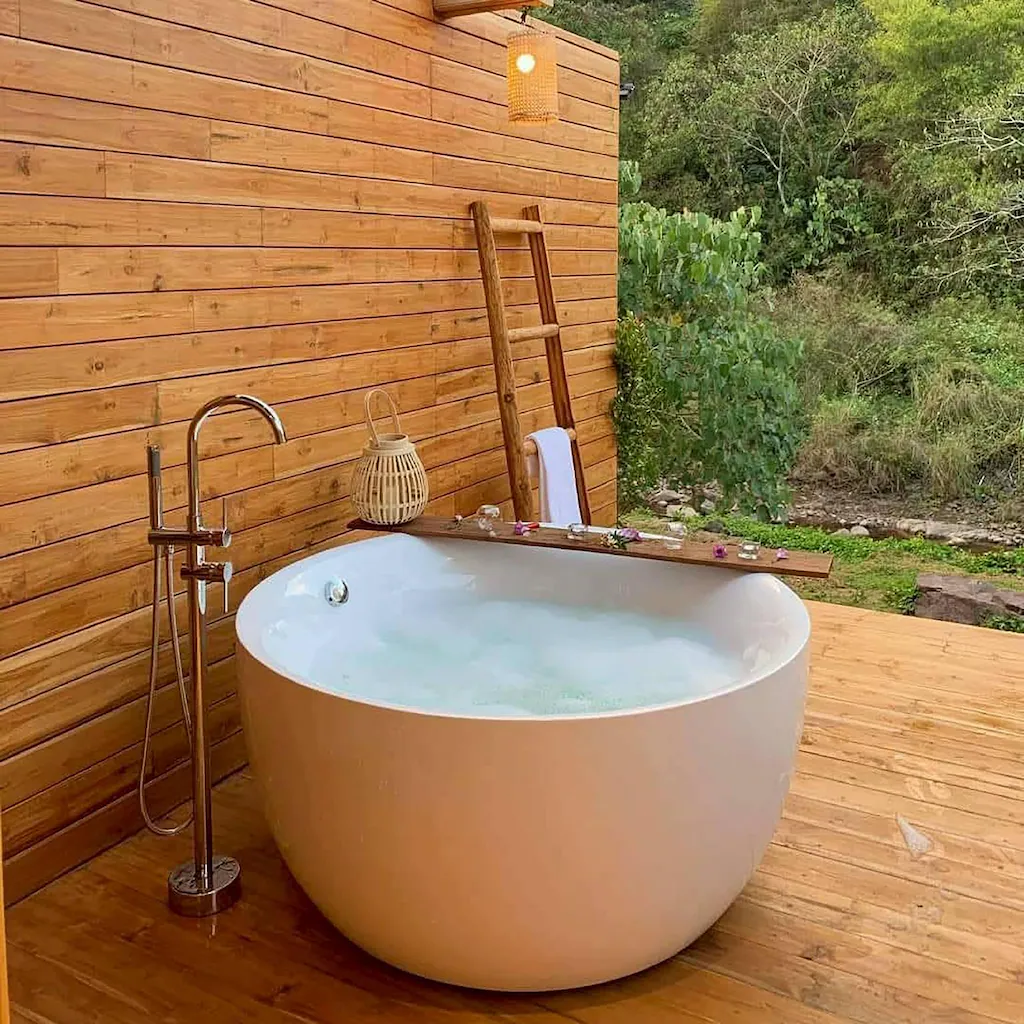 อ่างอาบน้ำทรงกลมสีขาวพร้อมน้ำไหลตั้งอยู่บนดาดฟ้าไม้ ล้อมรอบด้วยผนังไม้ บันได และพืชพรรณสีเขียวในฉากหลัง—เหมาะสำหรับการผ่อนคลายที่ ที่พักบ่อเกลือ.