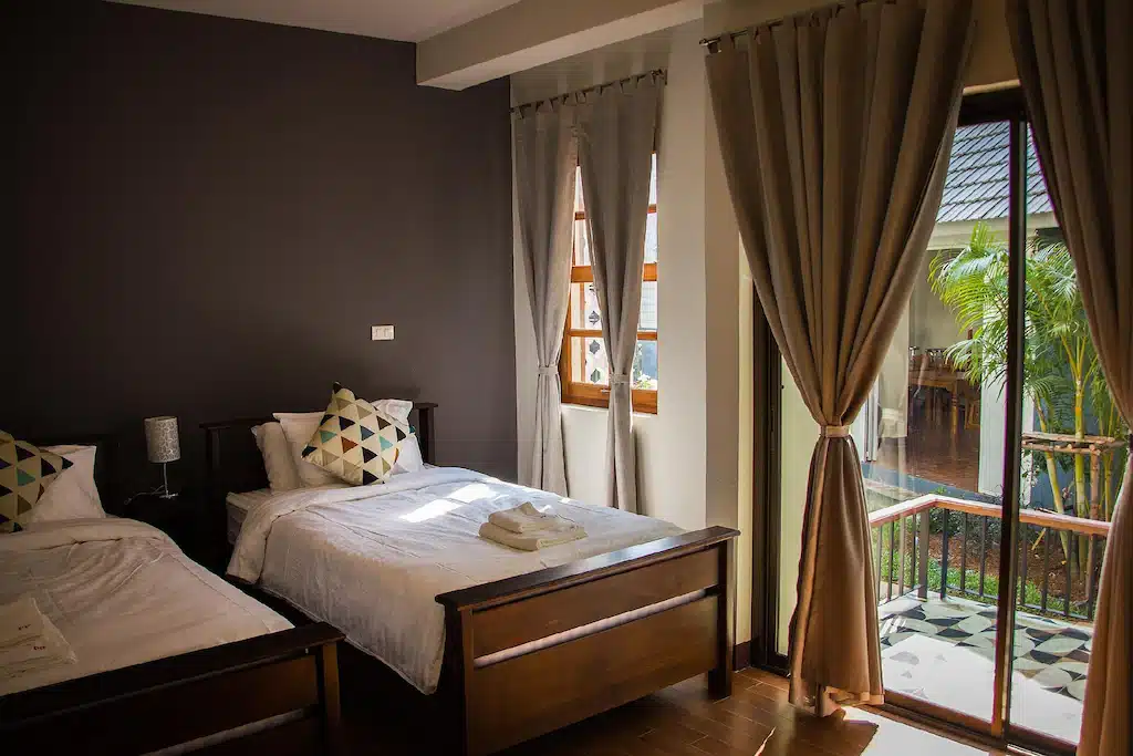 ห้องนอนที่อบอุ่นใน ที่พักบ่อเกลือ พร้อมเตียงเดี่ยวสองเตียง โคมไฟข้างเตียง ผ้าเช็ดตัวพับไว้ และหน้าต่างบานใหญ่พร้อมผ้าม่านที่มองเห็นระเบียงและสวนด้านนอก.