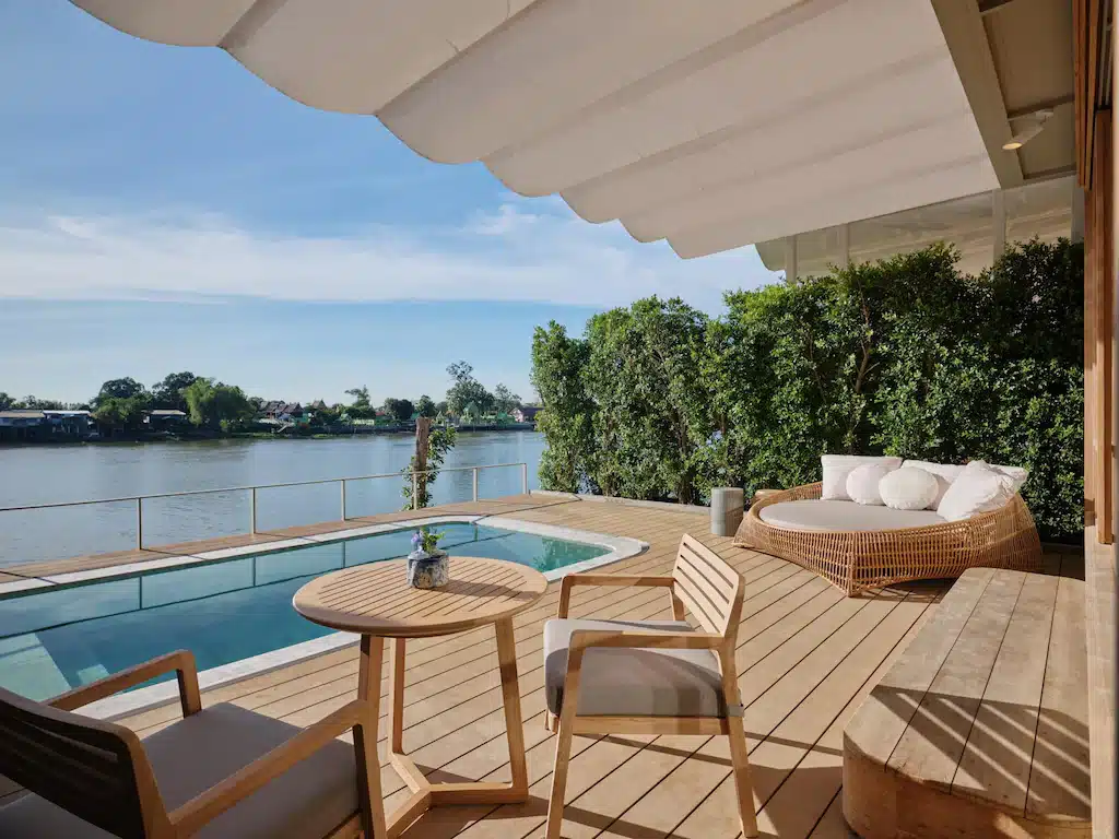 ระเบียงไม้พร้อมสระว่ายน้ำขนาดเล็ก เตียงนอนกลางวันทรงกลม และเฟอร์นิเจอร์ลานพักผ่อน มองเห็นแม่น้ำที่ที่พักริมน้ำอยุธยาอันเงียบสงบ รายล้อมไปด้วยธรรมชาติสีเขียวและท้องฟ้าสีครามเป็นฉากหลัง.